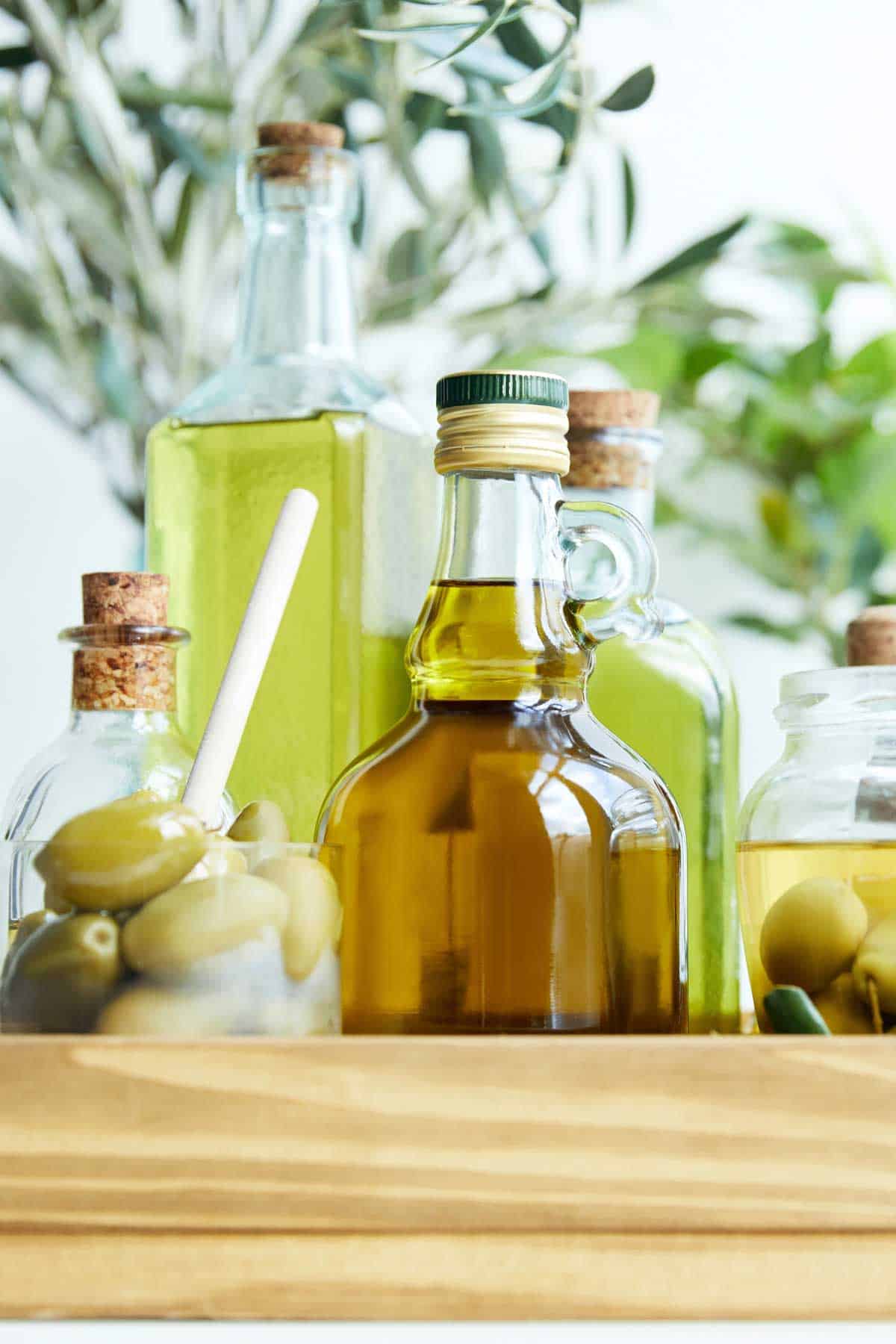 Several glass bottles filled with olive oil are arranged together, with jars of green olives and leafy olive branches in the background.
