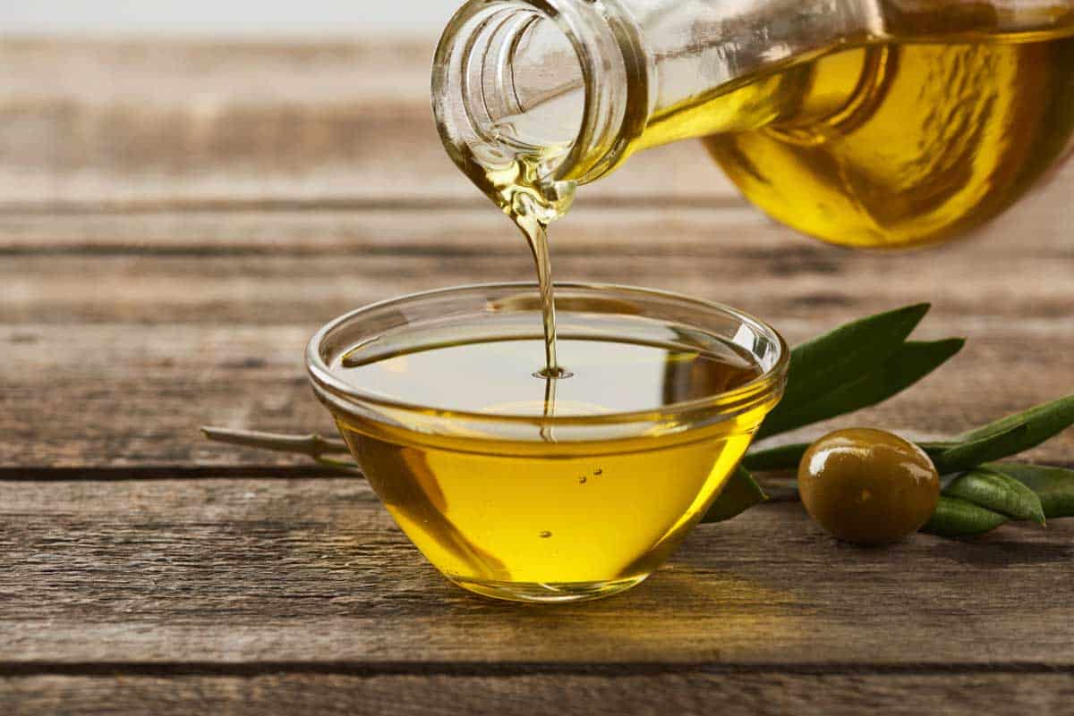 Olive oil is being poured from a glass bottle into a small glass bowl, with an olive and leaves on a rustic wooden surface.