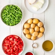 Bowls of halved cherry tomatoes, whole baby potatoes, chopped green beans, sliced onion, and various seasonings arranged on a white wooden surface.