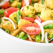 A bowl of potato salad with green beans, cherry tomatoes, and onion slices, mixed with herbs and dressing.