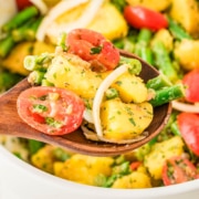 A wooden spoon holds a serving of potato salad with green beans, cherry tomatoes, onions, and herbs above a white bowl containing more of the salad.