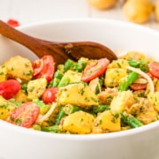 A white bowl filled with potato salad, green beans, cherry tomatoes, onions, and herbs, with a wooden spoon resting inside.