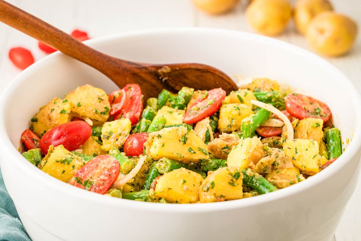 A white bowl filled with potato salad, green beans, cherry tomatoes, onions, and herbs, with a wooden spoon resting inside.