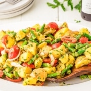 Oval white plate with potato salad, green beans, cherry tomatoes, onions, and herbs, with a wooden spoon in the dish and plates in the background.