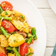 A plate of potato salad with green beans, grape tomatoes, onions, and herbs, served on a white dish next to a fork and a teal napkin.
