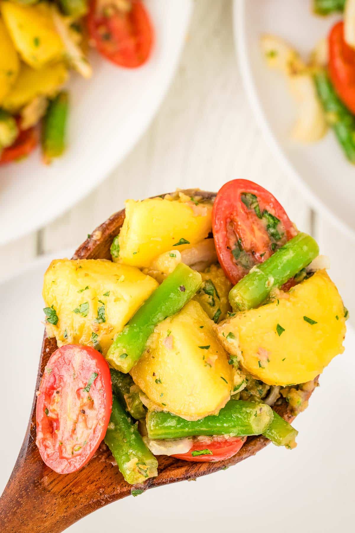 A wooden spoon holds a serving of potato salad with green beans, cherry tomatoes, onions, and herbs.