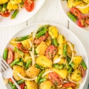 Plates of potato salad with green beans, cherry tomatoes, and onion slices are served on a white table, shown from above.
