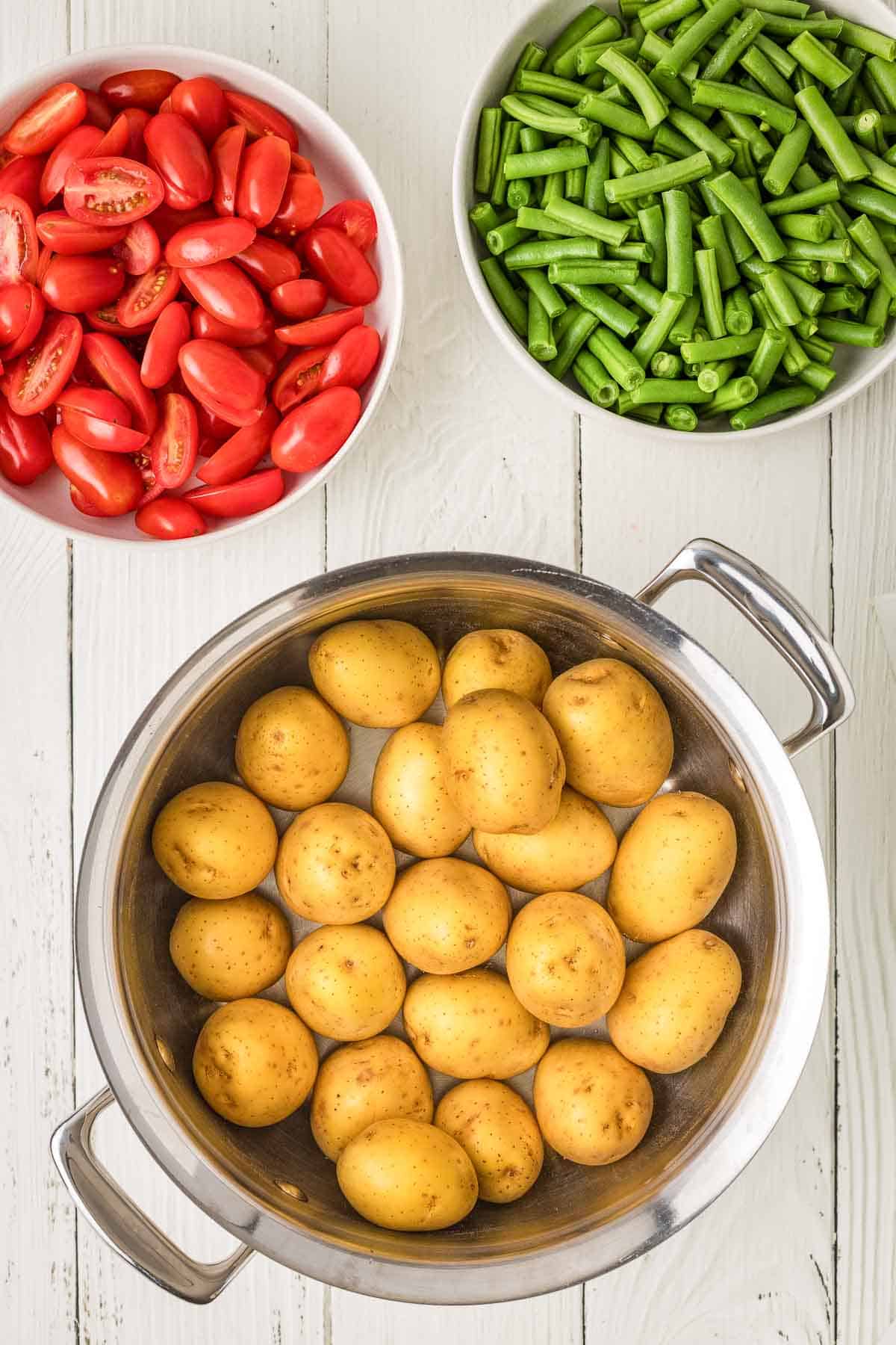 Three bowls on a white wooden surface: one with halved grape tomatoes, one with chopped green beans, and one with whole baby potatoes in a metal pot.