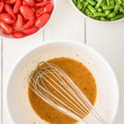 A metal whisk in a bowl of vinaigrette dressing, with bowls of chopped green beans and halved grape tomatoes on a white wooden surface.