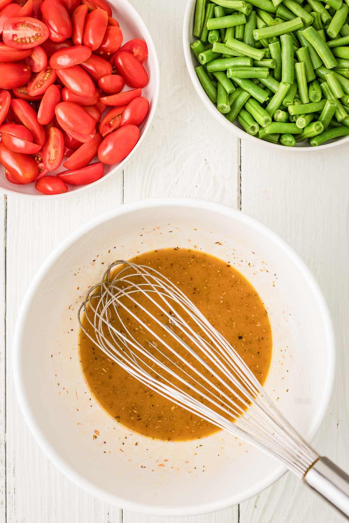 A metal whisk in a bowl of vinaigrette dressing, with bowls of chopped green beans and halved grape tomatoes on a white wooden surface.