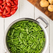 A pot of cut green beans in water sits on a white wooden surface, next to a bowl of halved grape tomatoes and a cutting board with whole yellow potatoes.