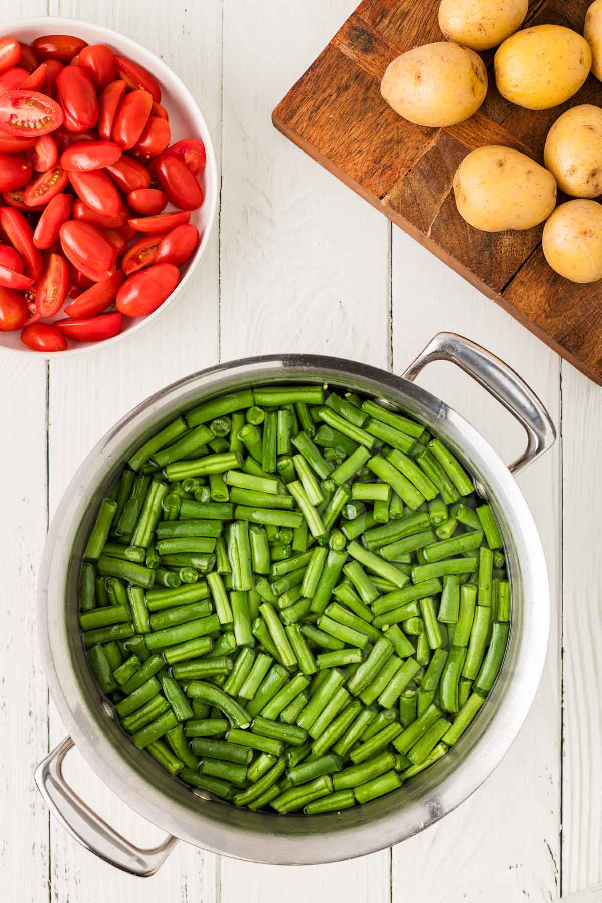 A pot of cut green beans in water sits on a white wooden surface, next to a bowl of halved grape tomatoes and a cutting board with whole yellow potatoes.