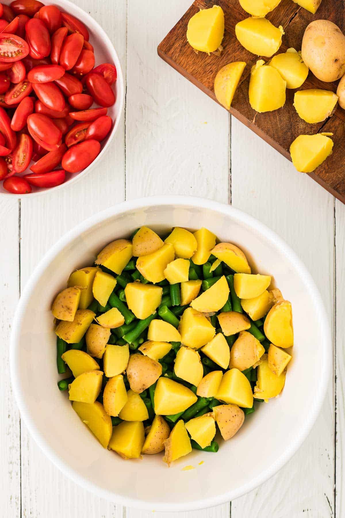 A white bowl with chopped yellow potatoes and green beans, a cutting board with more potatoes, and a bowl of halved cherry tomatoes on a white wooden surface.