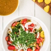 A white bowl filled with sliced tomatoes, onions, potatoes, and chopped herbs sits on a white table; a cutting board with potatoes and a bowl of broth are nearby.