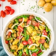 A bowl of potato salad with green beans, cherry tomatoes, onions, and herbs sits on a white table, surrounded by fresh potatoes, garlic, and tomatoes.