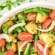 A bowl of potato salad with green beans, cherry tomatoes, sliced onions, and chopped herbs, with parsley scattered on a white wooden surface.