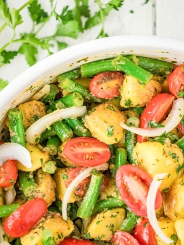A bowl of potato salad with green beans, cherry tomatoes, sliced onions, and chopped herbs, with parsley scattered on a white wooden surface.