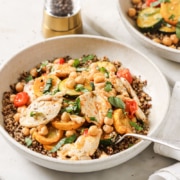 A bowl of quinoa topped with grilled chicken, chickpeas, sliced yellow squash, cherry tomatoes, and fresh herbs, with a fork resting on the side.