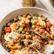 A bowl of quinoa salad with chickpeas, sliced chicken, cherry tomatoes, and fresh herbs, with a fork resting inside the bowl and a pepper grinder in the background.