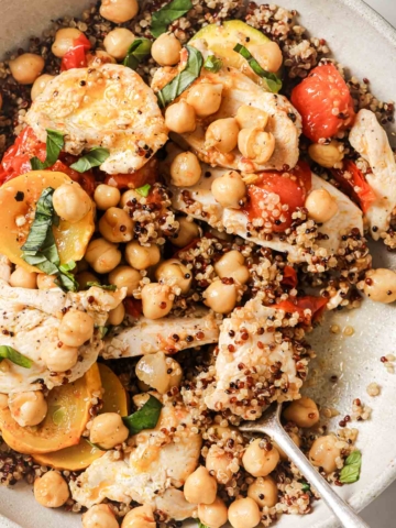 A bowl of quinoa salad with chickpeas, sliced chicken breast, cherry tomatoes, and fresh basil, with a fork resting on the side.