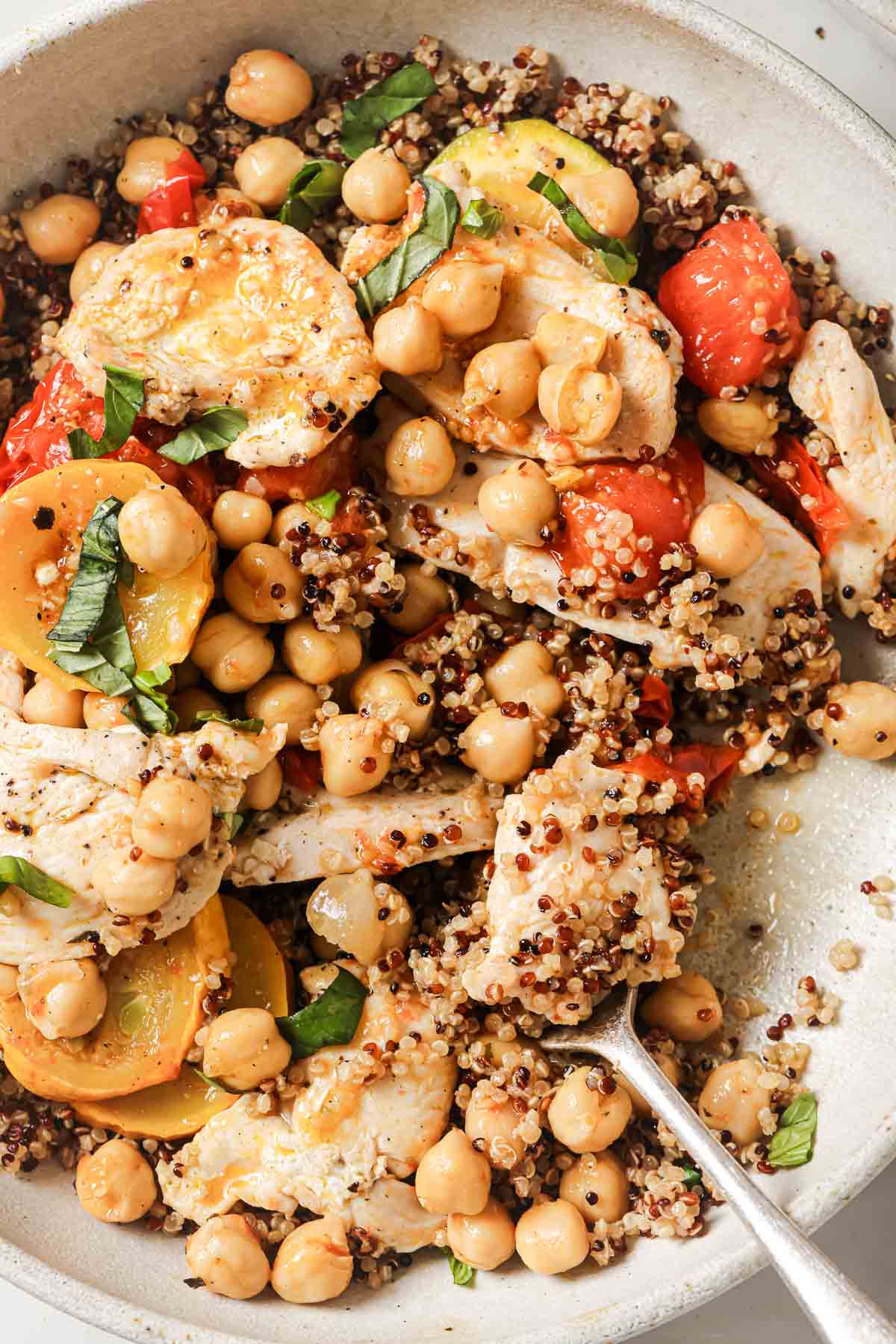 A bowl of quinoa salad with chickpeas, sliced chicken breast, cherry tomatoes, and fresh basil, with a fork resting on the side.