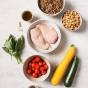 Overhead view of raw chicken breasts, cherry tomatoes, basil, garlic, yellow squash, zucchini, canned chickpeas, cooked quinoa, olive oil, salt, and pepper on a white surface.