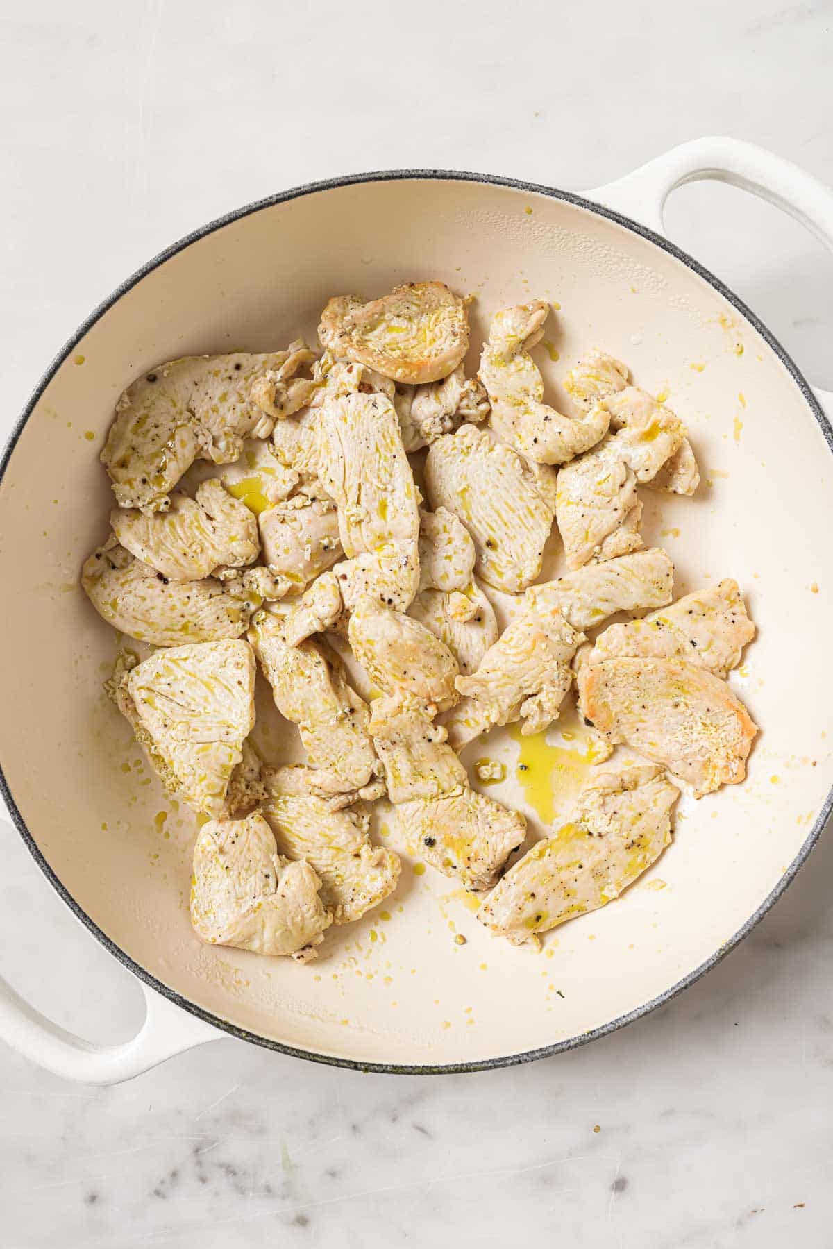 Cooked, seasoned pieces of chicken breast in a white Dutch oven on a marble surface.
