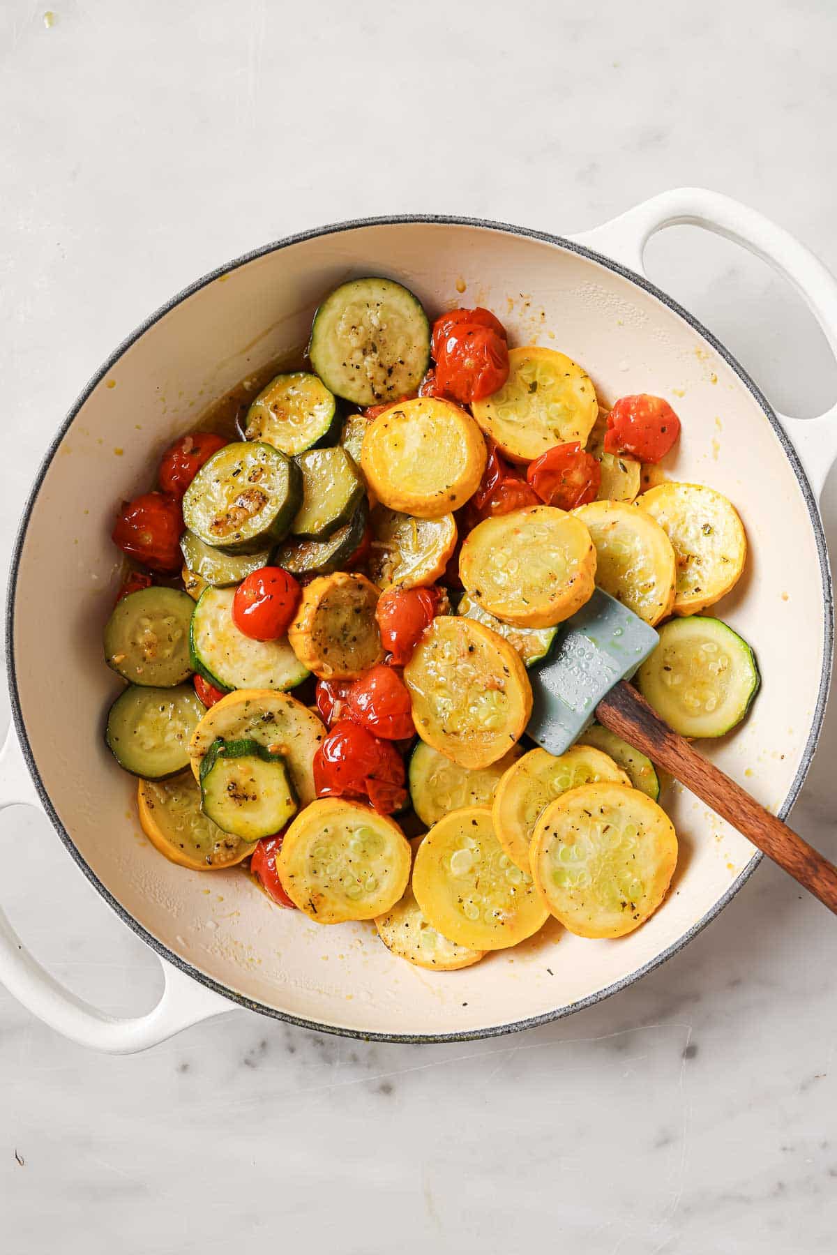 A white pot containing saut&eacute;ed yellow squash, zucchini, and cherry tomatoes, with a spatula resting inside on a marble surface.