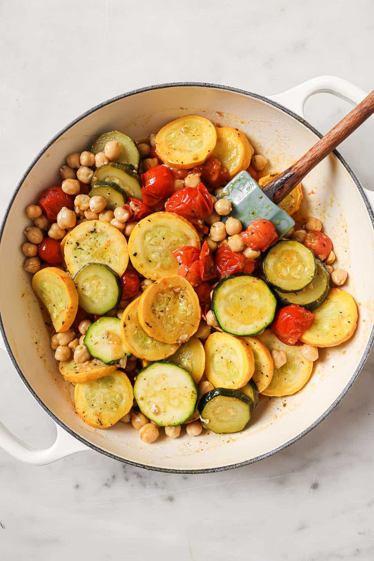 Saut&eacute;ed zucchini, yellow squash, cherry tomatoes, and chickpeas in a white pan with a spatula on a marble surface.