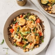 A bowl of quinoa topped with roasted vegetables, chickpeas, cherry tomatoes, and fresh herbs sits on a marble surface next to a pepper grinder and a small bowl of chopped greens.