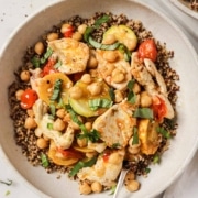 A bowl of quinoa topped with grilled chicken, chickpeas, zucchini, cherry tomatoes, and chopped herbs, with a fork resting in the bowl.