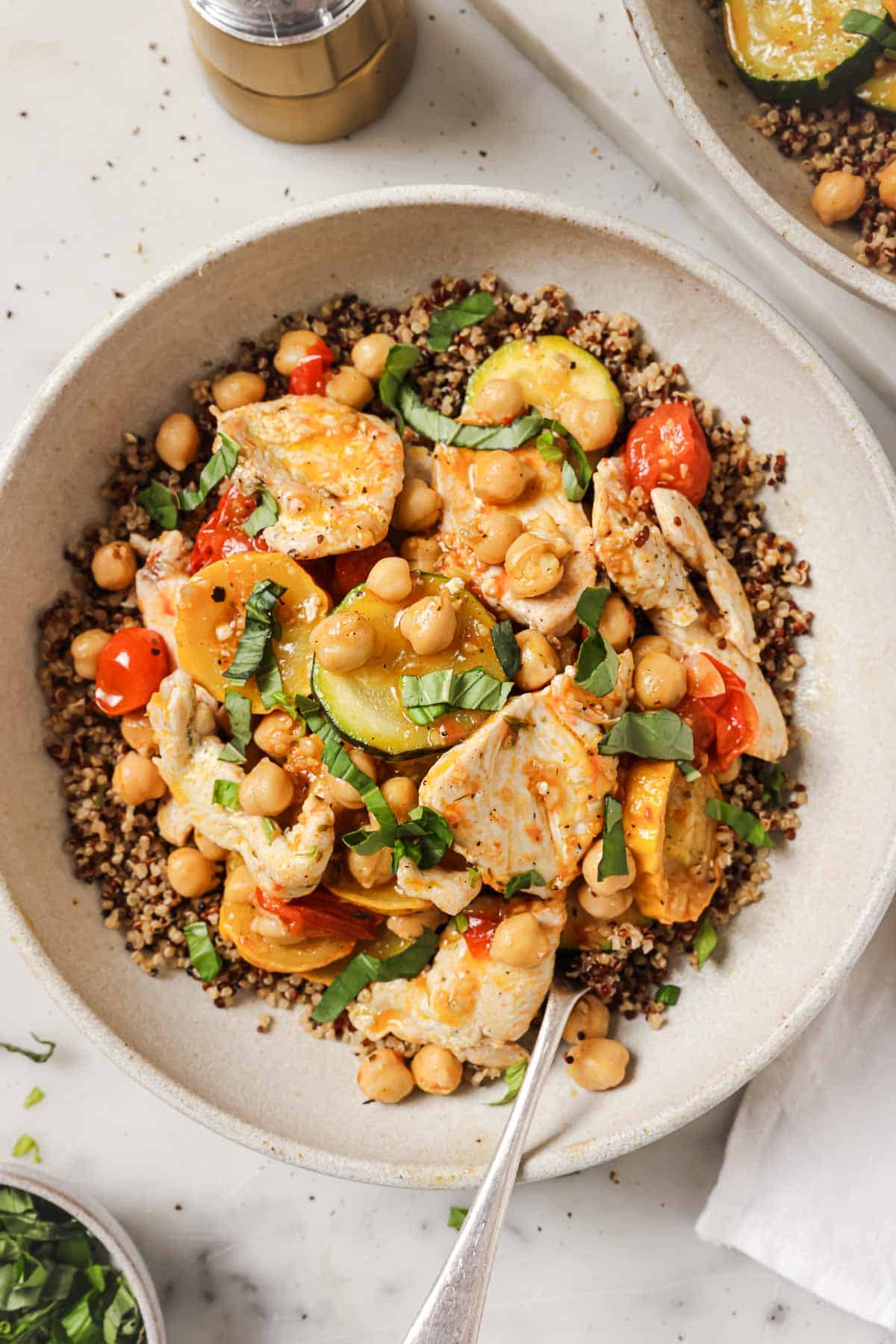 A bowl of quinoa topped with grilled chicken, chickpeas, zucchini, cherry tomatoes, and chopped herbs, with a fork resting in the bowl.