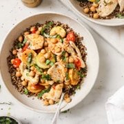 A bowl of quinoa topped with sliced grilled chicken, chickpeas, cherry tomatoes, cucumber, and fresh herbs, with a fork and a white napkin beside it.