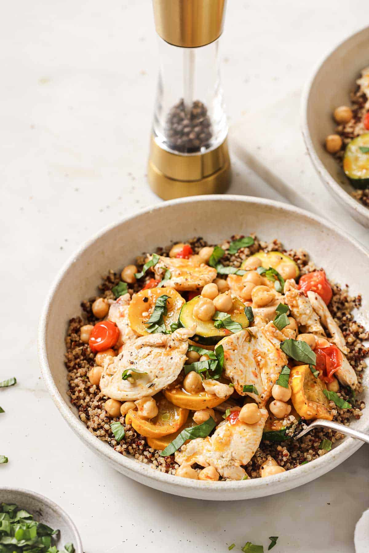 A bowl of cooked quinoa topped with sliced chicken breast, cherry tomatoes, chickpeas, zucchini, and fresh basil, with a pepper grinder in the background.