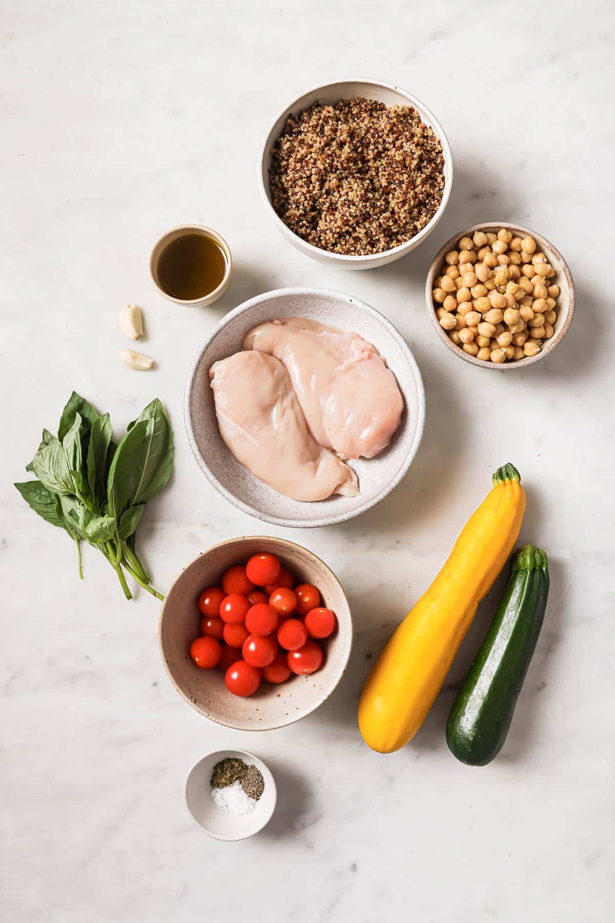 Overhead view of raw chicken breasts, cherry tomatoes, basil, garlic, yellow squash, zucchini, canned chickpeas, cooked quinoa, olive oil, salt, and pepper on a white surface.