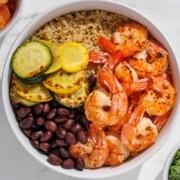 A bowl filled with cooked shrimp, black beans, grilled zucchini and squash, and quinoa, arranged in sections on a white surface.
