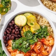 A bowl filled with shrimp, black beans, grilled zucchini, yellow squash, quinoa, and chopped greens, with a spoon and side bowl of sauce nearby.