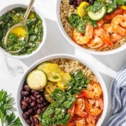Two bowls filled with quinoa, shrimp, black beans, zucchini slices, and green herb sauce, with extra sauce in a small bowl and parsley garnish on the side.