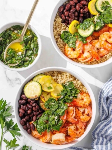 Two bowls filled with quinoa, shrimp, black beans, zucchini slices, and green herb sauce, with extra sauce in a small bowl and parsley garnish on the side.
