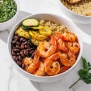 A bowl of cooked shrimp, black beans, and sliced grilled zucchini on quinoa, placed on a white surface with a green leafy herb nearby.