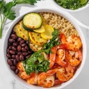 A bowl containing grilled shrimp, black beans, quinoa, zucchini, yellow squash, and fresh greens, arranged in separate sections on a white surface.