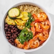 A bowl with quinoa, grilled zucchini and yellow squash, black beans, shrimp, and chopped greens on a marble surface.