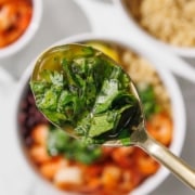 A close-up of a spoon holding chopped fresh herbs and oil, with a blurred bowl of food in the background.