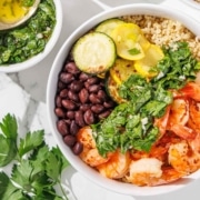 A bowl with shrimp, black beans, quinoa, grilled zucchini and squash, topped with green herb sauce. Fresh parsley and a bowl of sauce are nearby.