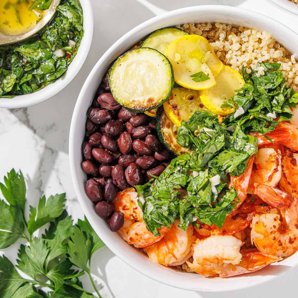 A bowl with shrimp, black beans, quinoa, grilled zucchini and squash, topped with green herb sauce. Fresh parsley and a bowl of sauce are nearby.