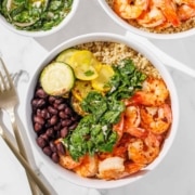A bowl with grilled shrimp, black beans, zucchini, yellow squash, chopped greens, and quinoa on a white surface with forks beside it.