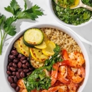 A bowl with shrimp, grilled zucchini, black beans, quinoa, and herbs next to a small bowl of green dressing with a spoon, on a white surface with parsley garnish.