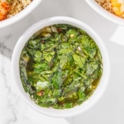 A white bowl filled with chimichurri sauce made from chopped herbs, garlic, and oil, placed on a white surface.