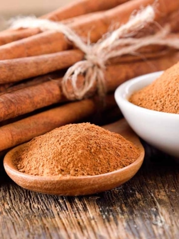 A bundle of cinnamon sticks tied with string is next to a small wooden spoon and a white bowl, both filled with ground cinnamon, on a rustic wooden surface.